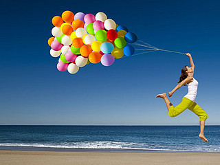 Eine junge Frau springt mit bunten Luftballonen über einen Strand.
