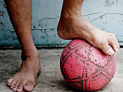 Zwei nackte Füße vor einer kaputten Hauswand; einer der Füße steht auf einem roten, abgenutzten Ball.