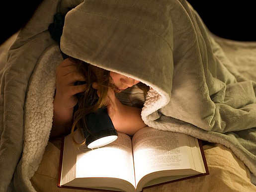 Ein junges Mädchen liegt unter der Decke und ließt mit ihrer Taschenlampe ein Buch