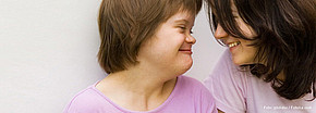 Ein Mädchen mit Down-Syndrom und eine Frau lächeln sich an und stecken ihre Köpfe zusammen