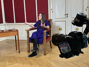 Eine Frau sitzt auf einem Stuhl vor rotem Hintergrund, im Vordergrund ist eine Kamera zu sehen