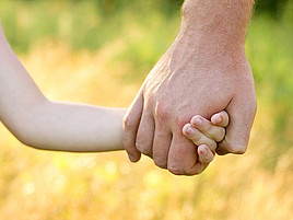 Die Hand eines Erwachsenen hält die Hand eines Kindes vor dem Hintergrund einer Wiese.