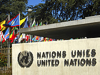 Informationstafel mit der Aufschrift "Nations unies" und "United Nations" sowie dem UN-Logo; im Hintergrund sind die Flaggen der UN-Mitgliedsstaaten, Bäume und blauer Himmel zu sehen.