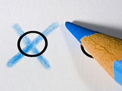 Ein blauer Buntstift malt ein Kreuz in einen schwarzen Kreis auf weißem Papier.