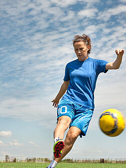 Eine junge Frau im blauen Dress kickt einen gelben Fußball auf einem Rasen unter einem leicht bewölkten Himmel.