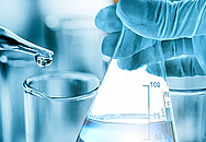 Das Bild zeigt eine Hand, die eine Flüssigkeit mittels einer Pipette in einem Labor in ein Reagenzglas tropft