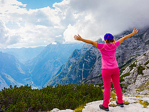Frau blickt von einem Aussichtspunkt auf das Bergpanaorama und breitet die Arme wie zur Umarmung aus