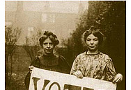 Historisches Bild zeigt zwei Frauen, die ein Plakat mit der Aufschrift votes for women steht.