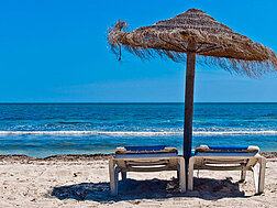 Zwei Liegestühle unter dem Sonnenschirm am Strand mit Blick auf das blaue Meer