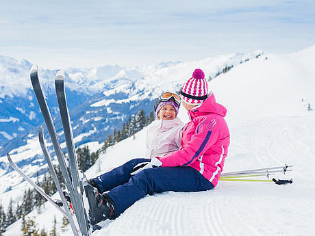 Zwei SkifahrerInnen sitzen auf einem Hügel, umgeben von winterlicher Landschaft 