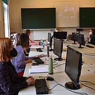 Einige SchülerInnen mit Masken sitzen im einem Raum, vor ihnen jeweils ein Computer 