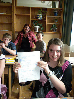 Die Schülerin hält ihren Test hoch; im Hintergrund sind weitere Kinder und die Lehrerin zu sehen.