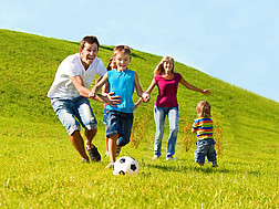 Eine Familie spielt auf einer Wiese unter blauem Himmel Fußball.