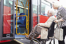 Ältere Frau schiebt älteren Mann im Rollstuhl über die Einstiegsrampe eines Busses