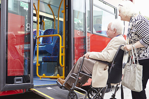 Ältere Frau schiebt älteren Mann im Rollstuhl über die Einstiegsrampe eines Busses