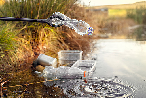 Plastikflaschen lagern am Flussrand und eine Zange greift entfernt eine Plastikflasche.
