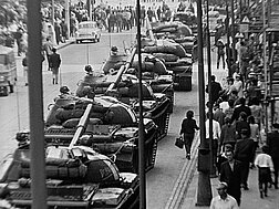 Panzer rollen eine Straße in Prag entlang, daneben Menschen auf dem Gehsteig, die unberührt vom Treiben ihrer Wege gehen