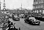 Historisches Foto zeigt eine Straße gesäumt von einer Menschenmenge, die die deutsche Wehrmacht in ihren Autos mit dem Hitler-Gruß empfängt.