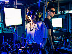 Zwei Forscher:innen mit Brille, in blauem Licht, vor Bildschirmen und Tischen mit technischen Geräten