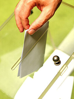 Eine Hand steckt einen zusammengefalteten, weißen Stimmzettel in eine durchsichtige Wahlurne.