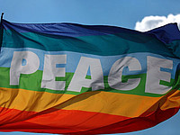 Eine wehende Fahne in den Regenbogen-Farben mit der Aufschrift "Peace" vor einem blauen Himmel mit einer Wolke