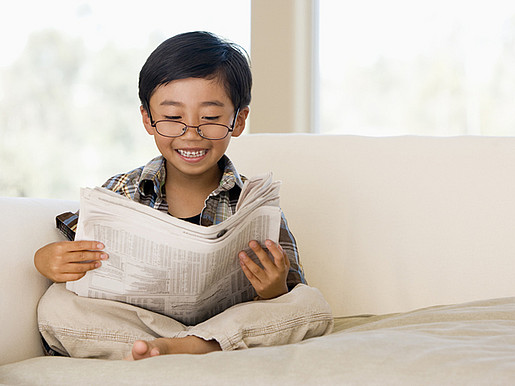 Ein Junge mit Brille sitzt auf einem Sofa und liest Zeitung.