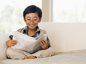 Junge sitzt im Schneidersitz auf einer Couch und liest Zeitung