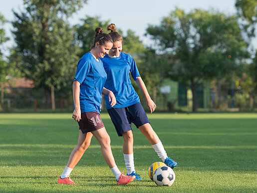 Zwei junge Frauen im Fußballdress trainieren auf einem Rasen; im Hintergrund sind Bäume zu sehen.