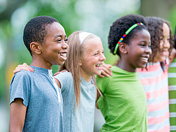 Das Bild zeigt Kinder verschiedener Hautfarbe, die Arm im Arm lächelnd darstehen