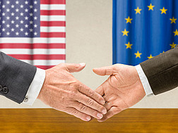 Das Bild zeigt zwei Menschen, die sich die Hand reichen. Im Hintergrund die Flaggen der USA und der Europäischen Union 