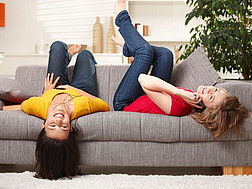 Zwei junge Frauen liegen lachend verkehrt auf einem Sofa. Eine von ihnen telefoniert.