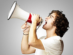 Ein junger Mann im T-Shirt vor grauem Hintergrund ruft etwas durch ein Megaphon, das er in seinen Händen hält.