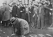 Eine Menschenmenge umringt einen jüdischen Mann, der knieend die Straße putzen muss.