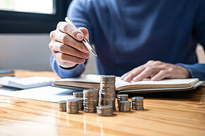 Ein Stapel Geldmünzen liegt auf einem Tisch. Eine Person sitzt mit Stift in der Hand vor einem Notizbuch.