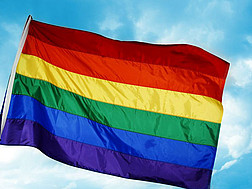 Eine Flagge in den Farben des Regenbogens weht vor einem leicht bewölkten Himmel im Wind.